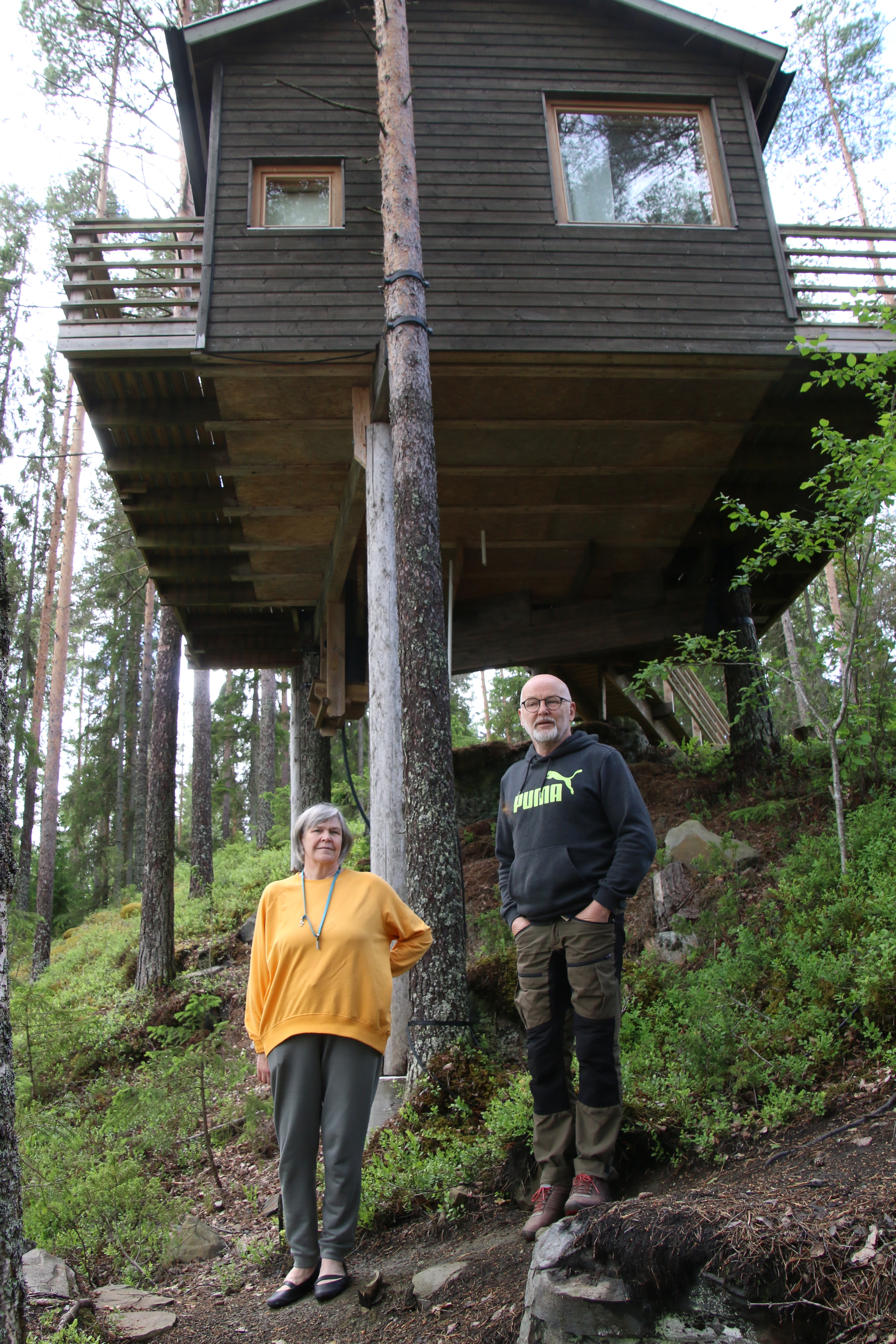 Kuva, jossa kaksi henkilöä seisoo metsässä. Taustalla puiden varaan rakennettu mökki.
