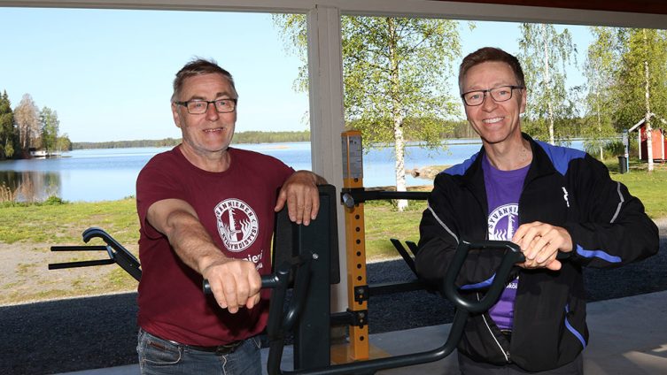 Kaksi miestä (Jari Partanen ja Pekka Kääriäinen) hymyilee kuntoilulaitteiden vieressä, taustalla kaunis kesäinen järvimaisema.