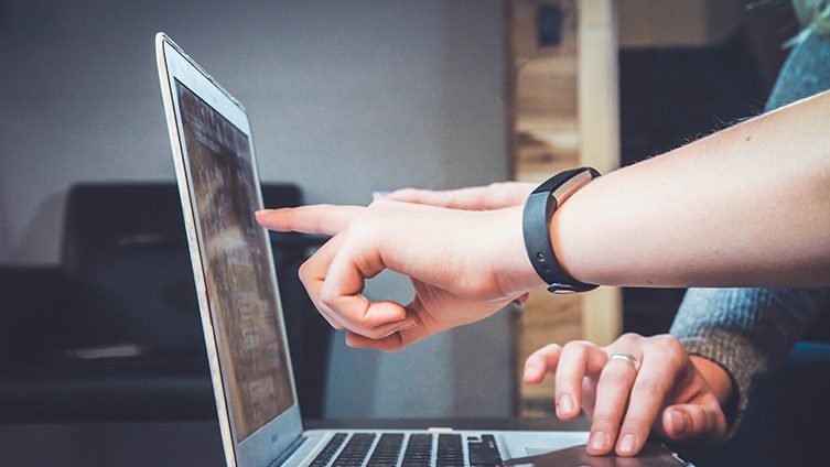 Kannettava tietokone, jonka ruutua osoittaa käsi, toisen henkilön kädet käyttävät näppäimistöä