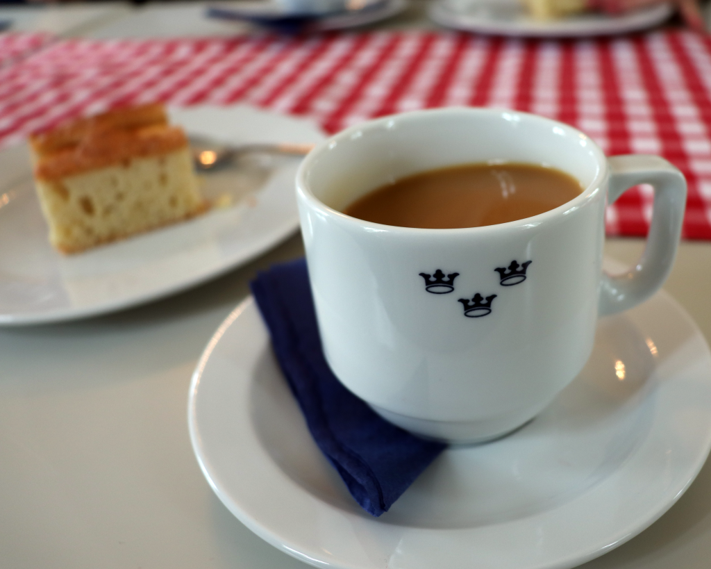 Kuva kahvikupista, jossa on kolme kruunua. Taustalla piirakanpala.